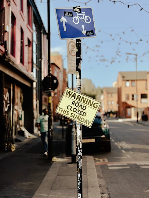 A street scene in Spitalfields featuring a metal pole with two signs attached; the upper sign is a blue and white cycling lane sign indicating one-way traffic for bicycles and pedestrians, with an arrow pointing upward and downward, and the lower sign is a yellow and black warning notice reading 'Road Closed This Sunday.' The pole is positioned on the sidewalk, with the background showing a row of brick buildings, shopfronts, and a parked black van. The scene is lit by natural daylight, and the overall setting reflects an urban environment prepared for possible road closures affecting residential and commercial moving activities, often managed by local removals services such as Man with Van Spitalfield, ensuring efficient home relocation and furniture transport during restricted access periods.