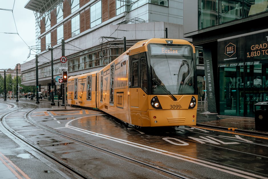 A yellow tram with the number 3099A travels along wet railway tracks on a city street, passing modern office buildings and retail stores. The tram is positioned on the curved track with visible overhead power cables above it. Nearby, traffic signals are illuminated, and the street surface appears damp, indicating recent rain. Adjacent to the tram, there is a sidewalk and a glass-fronted commercial building displaying signs. This scene reflects urban transportation infrastructure associated with house removals and moving logistics, highlighting the importance of efficient travel routes in central London, such as from Liverpool Street Station to E1 homes, as managed by Man with Van Spitalfield.