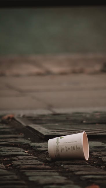 A white paper cup with green text and logo, discarded on a weathered wooden doorstep with visible grime and dirt, lying on its side. The door is partially open, revealing the entrance to a house with a green painted wall in the background. The scene is outdoors during daylight with natural lighting. This image shows waste materials possibly collected after a house move or packing process, relevant to home relocation and packing and moving services provided by Man with Van Spitalfield. The cup's placement suggests it may be part of disposal or waste collection efforts during a furniture transport or removals operation, aligning with guidance on waste disposal after moving in the Tower Hamlets area.