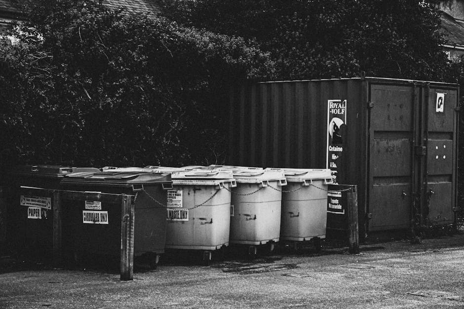 A black and white photograph showing a row of waste disposal containers positioned outside a residential building, with a large metal storage shipping container behind them. The waste bins include a dark-colored recycling bin labeled 'PAPERS,' and four larger white bins marked 'WASTE ONLY,' all placed on a paved surface next to a wooden post. The containers are arranged near the entrance of a property, likely in a driveway or designated waste area, with trees or foliage visible in the background. The scene depicts an outdoor setting related to home relocation and moving logistics, where disposal of waste materials and packing debris could be part of the house removal process. The image highlights the importance of proper waste management during the packing and loading stages handled by companies like Man with Van Spitalfield, especially after a house move or furniture transport involving packing and unpacking of various items.
