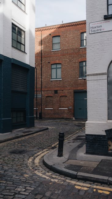 An outdoor scene at the corner of a narrow cobblestone street in Spitalfields during daylight, with two modern buildings on either side. The left building has a white facade with large windows, while the right has a white brick wall displaying a street sign reading 'Hoxton Square.' In the background, an older red brick building features multiple windows with grey frames and closed shutters. The street is bounded by short black bollards, and the pavement shows signs of recent activity related to home relocation or furniture transport. This setting reflects a typical urban environment for house removals or loading during a property move, with no visible vehicles or moving equipment present.