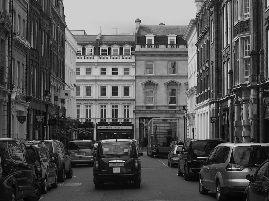 Black and white photograph of a narrow urban street scene in Spitalfields, showing parked cars lining both sides of the street in front of historic multi-storey buildings with ornate facades and large windows. In the background, a delivery van with its back doors open is positioned near the curb, suggesting a loading or unloading activity related to home relocation or furniture transport. The scene captures the exterior environment where a professional removals service, such as Man with Van Spitalfield, may be engaged in packing and moving processes. The street features typical architectural details of the area, with windows, balconies, and decorative elements, and appears to be during daytime with natural lighting, emphasizing the urban context for house removals and tight parking considerations.