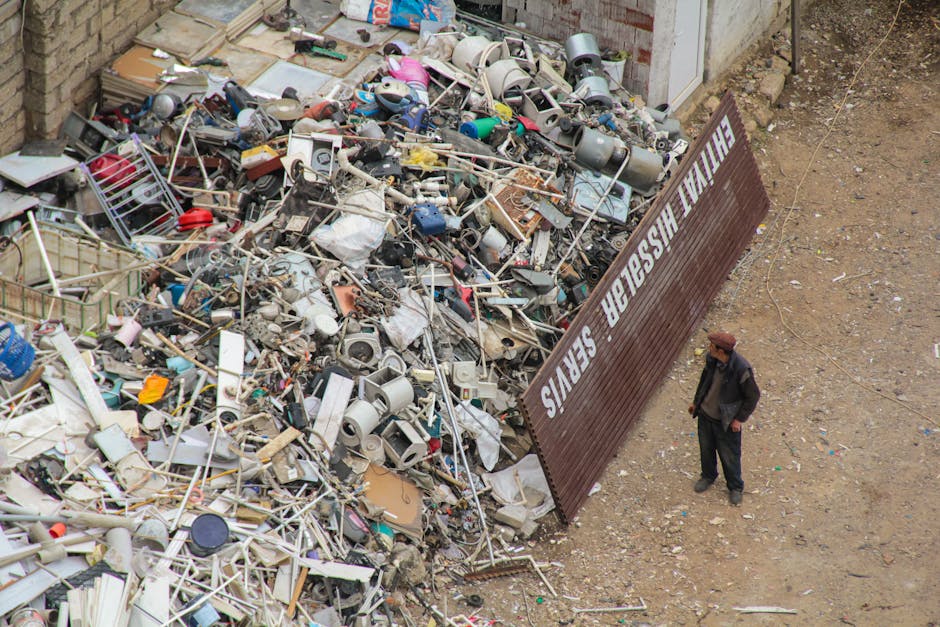 An outdoor scene showing a large pile of assorted household waste, including broken electronics, plastic containers, metal objects, and miscellaneous debris, situated next to a brown metal fence with white lettering that reads 'MAN WITH VAN REMOVALS SERVICES.' The waste heap appears to be in a designated disposal area, with some materials spilled onto the bare, uneven ground. A man wearing dark clothing and a brown hat is standing nearby, observing the area from the right side of the image. The background includes a portion of brick wall and a doorway, indicating this scene is adjacent to a residential or commercial property awaiting waste collection or home relocation clearance. The setting is well-lit by natural daylight, illustrating the typical clutter involved in house removals or bulky waste disposal, similar to services offered by [COMPANY_NAME] in the context of professional packing, loading, and transport of household items.