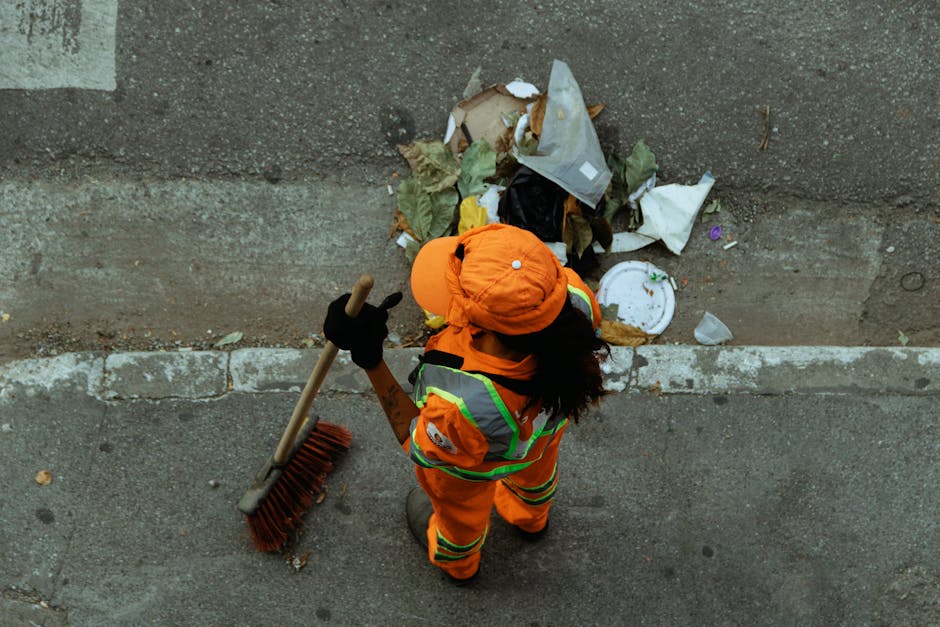 A person dressed in an orange high-visibility jacket, matching cap, and black gloves, is standing on a pavement next to a wall, holding a leaf rake with a wooden handle and red bristles. The individual appears to be involved in clearing debris and waste materials from the ground, which includes cardboard, plastic wrappers, paper plates, and other household rubbish. The waste is gathered along the edge of the pavement near the wall, indicating a clean-up or waste removal process. The scene captures a moment of manual waste collection, possibly as part of a house removal or space clearing process, with the person preparing to gather and dispose of rubbish. This activity is related to logistics and proper handling of household waste in the context of home relocation or clearance, and the environment is well-lit with no visible vehicles or additional people in the frame. Man with Van Spitalfield specializes in removals and helping clients manage waste disposal efficiently during moves.