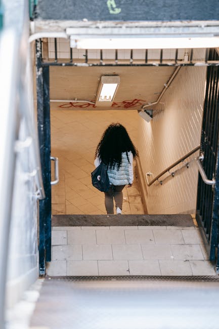 A person with long, curly dark hair, wearing a light-colored puffer jacket, grey trousers, and carrying a dark backpack, is descending a staircase inside a residential building. The staircase has wood