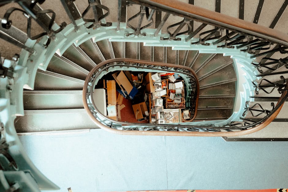 A person with long, curly dark hair, wearing a light-colored puffer jacket, grey trousers, and carrying a dark backpack, is descending a staircase inside a residential building. The staircase has wood