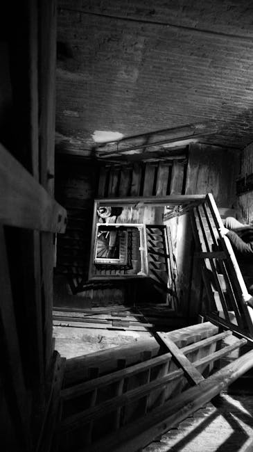 Black and white photograph showing a narrow, steep staircase inside a Victorian house, with wooden bannisters and handrails on both sides. The stairs are cluttered with various moving boxes, some wrapped in plastic and others in cardboard, along with furniture pieces including a wooden chair and parts of a small table. The scene is illuminated by natural light coming from an unseen source, casting shadows on the wooden floor and staircase. The image captures a moment during a home relocation process, with visible packing materials and furniture ready for transport, possibly by Man with Van Spitalfield, operating within the context of a house move involving careful navigation of tight interior spaces.