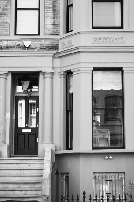 A black and white photograph of the front façade of a narrow Victorian terraced house with a decorative cornice and brickwork. The entryway features a small set of stone steps leading up to a black front door with a glass panel, surrounded by a white rendered frame. To the right of the door, there are two large, vertically oriented sash windows with dark frames, one of which has a partially visible roller blind inside. The ground level includes a small wrought iron fence and gate, with a paved area in front. The image captures the exterior architecture typical of London’s historic terraced housing, emphasizing the narrow frontage and tall, multi-paned windows, which pose specific challenges for house removals and furniture transport. This setting is representative of the type of property that Man with Van Spitalfield specializes in for safe and efficient home relocations and moving services.