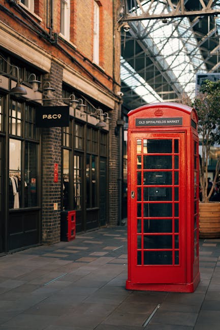 A traditional red British telephone kiosk with a rectangular shape and glass-paneled doors is situated on a paved sidewalk under an industrial-style glass and metal overhead structure. The kiosk displays the sign 'OLD SPITALFIELDS MARKET' at the top. To the left of the kiosk, there are brick building facades featuring large windows with black frames, and a black sign with the white text 'P&C9' mounted above a storefront entrance. The surrounding area includes wooden planters and other street furniture, with natural light illuminating the scene. The image captures a typical street scene in Spitalfields, London, emphasizing urban architecture and street furniture relevant to home relocation and moving logistics, as managed by companies like Man with Van Spitalfield during furniture transport and packing activities.