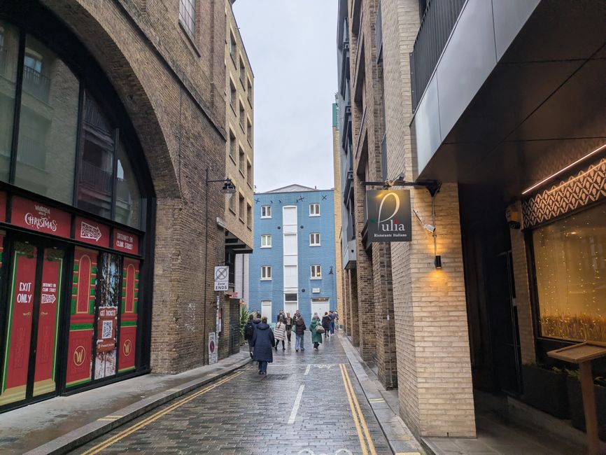 A narrow urban street scene showing a group of people walking along a wet cobblestone pavement between tall brick and modern commercial buildings. On the left, there is a storefront decorated with Christmas-themed window graphics and a sign indicating 'EXIT ONLY.' On the right, a business sign for 'Ulia Ristorante Italiano' is affixed to the brick facade, with external lighting illuminating its exterior. The street is lined with double yellow lines, and a traffic sign on the corner restricts vehicle access. In the background, a light blue residential building with white-framed windows rises into an overcast sky. The scene captures a typical city centre environment that could be involved in a home relocation or moving process, with pedestrians possibly en route to or from a nearby apartment or office, and the surrounding architecture indicating a densely built area suitable for furniture transport and packing activities in conjunction with services by Man with Van Spitalfield.