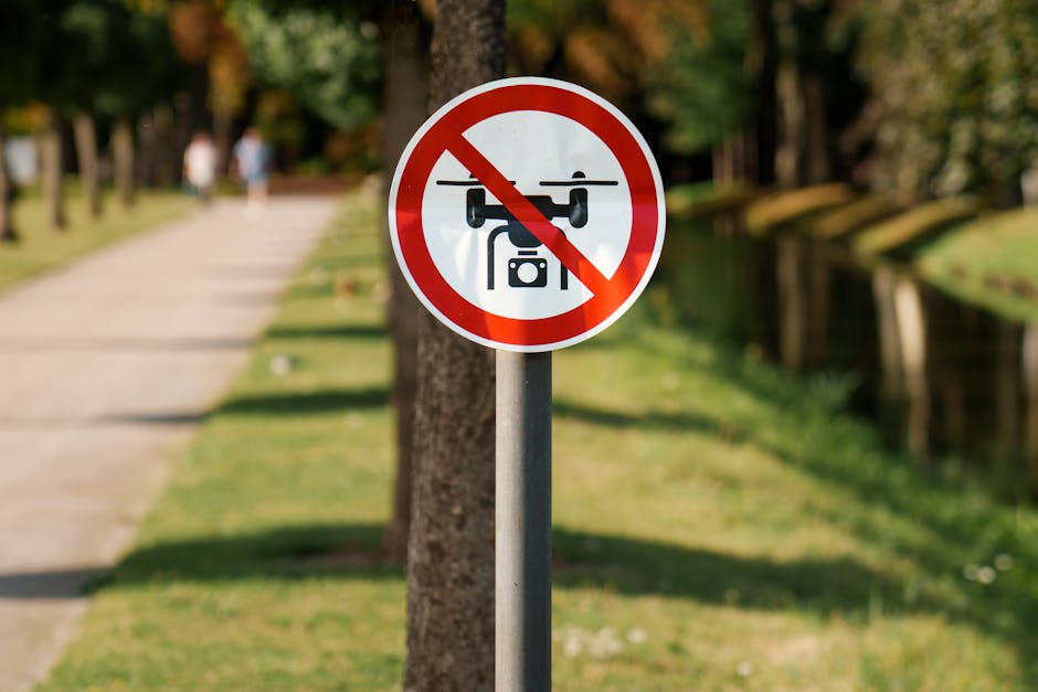 Outdoor scene featuring a round sign attached to a metal pole on a pathway beside a canal or small river, with a red border and a black drone graphic crossed out by a red line, indicating no drone flying permitted. The pathway is lined with trees and a grassy area, with three blurred figures walking in the distance. The setting appears to be during daylight with bright, natural lighting, and the background includes trees and a green landscape. This image relates to house removals or moving logistics, showing the importance of respecting local regulations for airspace when transporting or relocating items around residential or urban areas, and occasionally supported by companies like Man with Van Spitalfield specializing in removals in Tower Hamlets.