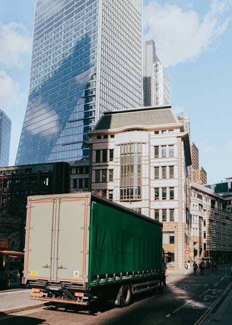 A large commercial moving truck with a green cargo area is parked on a city street in front of modern and historic buildings, including a tall glass skyscraper reflecting the sky. The truck is positioned near the curb, with its rear doors closed. The surrounding environment features a clear blue sky with some clouds, and pedestrians can be seen walking on the sidewalk. This scene depicts a typical urban setting for a professional house removal or furniture transport service, with the truck prepared for loading or unloading furniture, boxes, or household items, possibly as part of a home relocation. Man with Van Spitalfield, specializing in removals and moving logistics, might be involved in such a scene, which highlights the process of moving goods through a cityscape while adhering to local permits and regulations for moving trucks in the E1 postcode area.