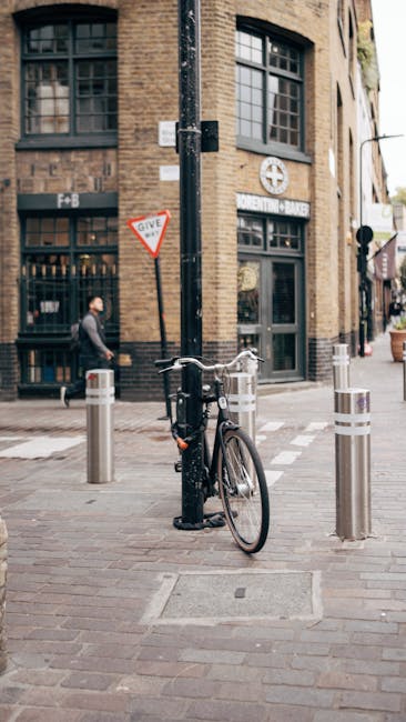The image shows a black bicycle locked to a metal pole on a city sidewalk, adjacent to four short, cylindrical bollards with reflective bands. Behind the bicycle, a pedestrian dressed in dark clothing is walking past a brick building with large windows, which has a sign indicating a financial institution. The building's exterior features arched windows and a sign for a local bank. The scene is well-lit with natural daylight, and the street is clean and organized, reflecting typical urban surroundings for house removals and packing and moving logistics, as seen in professional relocation services like those offered by Man with Van Spitalfield.