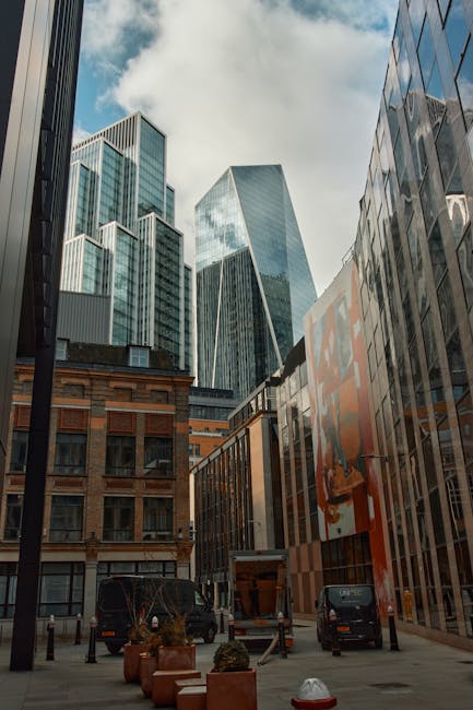 A view of a city street scene during daytime, featuring modern glass skyscrapers towering over older brick buildings. In the foreground, several black vans are parked on the pavement, with one van's rear door open, revealing packing materials such as cardboard boxes and plastic wrapping inside. The loading area appears to be used for home relocation or furniture transport, with visible equipment like trolleys and straps on the ground. Potted plants and bollards are positioned along the pavement, and the sky is partly cloudy with patches of blue. The scene captures the typical environment for a professional removals company like Man with Van Spitalfield, engaged in loading or unloading household belongings during a move in the central London area. The image emphasizes urban logistics and the process of preparing furniture and boxes for a home removals service.
